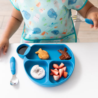 A child in an ocean-themed bib eats breakfast from a blue divided plate with Bumkins Spoon + Fork: Dark Blue at a white table.