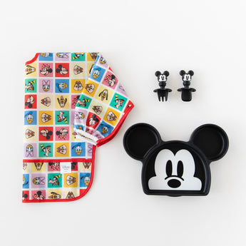 Bumkins Silicone Grip Dish: Mickey Mouse lays next to matching toddler utensils and a colorful Disney patterned bib on a white background.