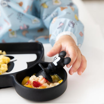 Close-up of a child's hand reaching for Bumkins Silicone Chewtensils®: Mickey Mouse on a Mickey-shaped plate with fruit.