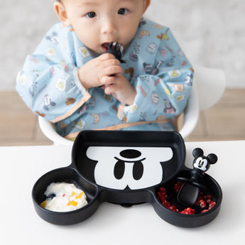 A baby in a blue bib chews a toddler utensil near Bumkins Silicone Grip Dish: Mickey Mouse with fruit & yogurt on a table.