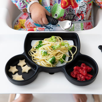 A toddler in a high chair enjoys self-feeding broccoli, pasta, and raspberries from Bumkins Essential Mealtime Set, Mickey Mouse.