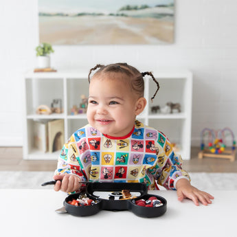 A girl in a colorful bib smiles as she eats from Bumkins Silicone Grip Dish: Mickey Mouse in a bright playroom with toys on shelves.