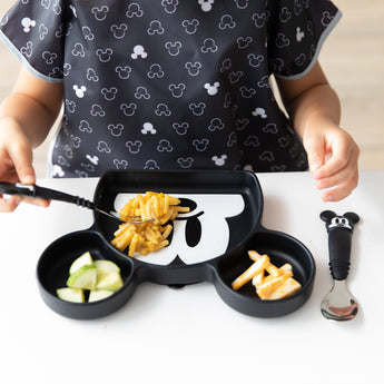 A toddler uses Bumkins Mickey Mouse Essential Mealtime Set to eat pasta and cucumbers at a white table.