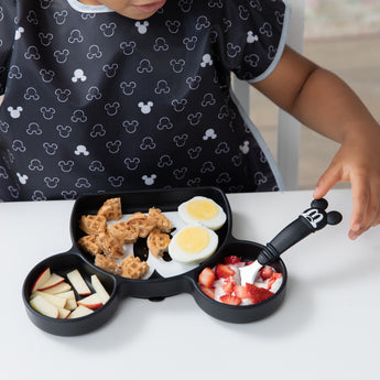 A toddler eats eggs, waffles, apples, and strawberries with Bumkins Mickey Mouse Essential Mealtime Set at a white table.