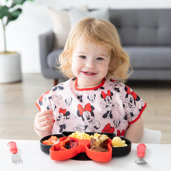 A young girl in a Bumkins Junior Bib: Minnie Mouse Classic smiles at a table with a Minnie Mouse-shaped plate of food and red utensils.