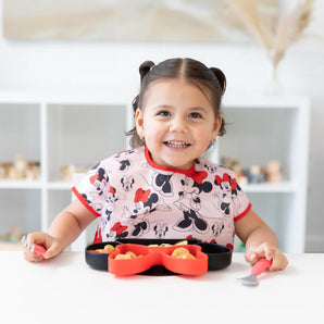 A young girl in pigtails smiles, wearing a Bumkins Junior Bib: Minnie Mouse Classic and holding utensils by a Minnie Mouse-shaped plate.