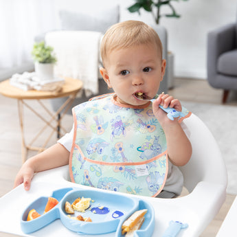 A boy in a pastel bib uses a fork to eat eggs and orange slices from a Bumkins Silicone Grip Dish: Stitch in a cozy living room.