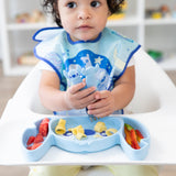 A young girl in a blue bib holds utensils over a Bumkins Silicone Grip Dish: Stitch with pasta, veggies, & fruit on a high chair tray.