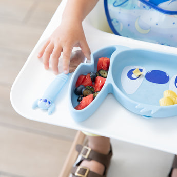 Close-up of a child's hand grabbing a spoon from Bumkins Spoon + Fork: Stitch next to a Stitch-shaped dish with fruit on a high chair tray.