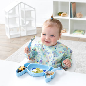 A young girl smiles at a white table, eating a meal with Bumkins Grip Dish Essential 3-Piece Mealtime Set: Disney Stitch.