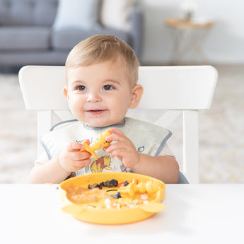 A boy sits at a white table, eating breakfast with Bumkins Grip Dish Essential Infant 3-Piece Mealtime Set: Disney Winnie the Pooh.