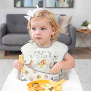 A girl in a bow  in a high chair holds a Bumkins Spoon + Fork: Winnie the Pooh by a plate of breakfast food,reaching into her bib pocket.