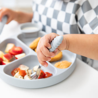 Close-up of a child in a checkered bib eating fruit from a Bumkins Silicone Grip Dish: Gray with a fork at a white table.