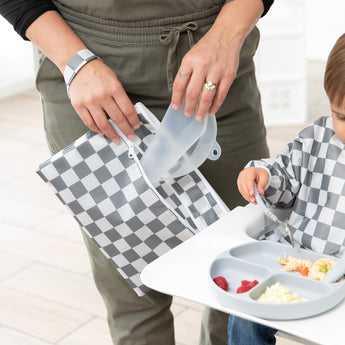 A mom and child use Bumkins On-The-Go Mealtime 6-Piece Set: Charcoal Check + Grey at mealtime in a bright room.