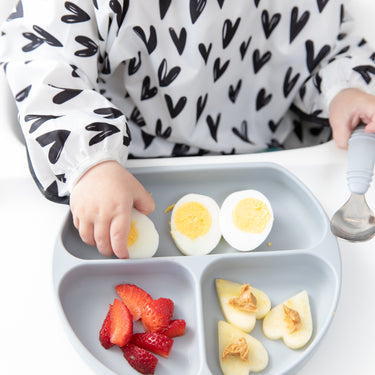 Close-up of a child in a bib with black hearts eating eggs and fruit with Bumkins Grip Dish Essential Toddler 4-Piece Mealtime Set: Gray.