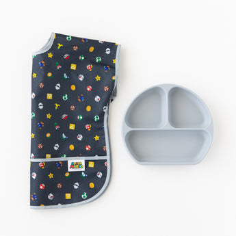 Bumkins Junior Bib: Super Mario™ Lineup  lays folded on a white background beside a gray silicone divided plate.