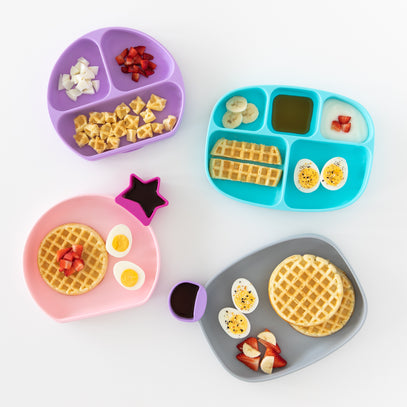 Four colorful divided plates with waffles, sliced fruits, and boiled eggs. Plates are pink, purple, blue, and gray, each with a small separate section holding syrup or sauce containers in fun shapes.