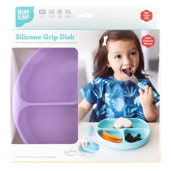A child smiles while eating from a turquoise Bumkins silicone grip dish, packaged with the lavender version featuring a suction base.
