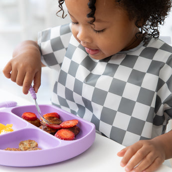 Close-up of a girl in a gray & white bib eating fruit from a purple plate with Bumkins Spoon + Fork: Lavender.