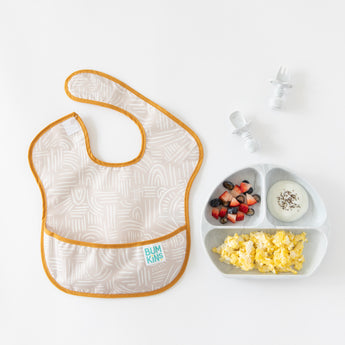 Bumkins Grip Dish Essential Infant 4-Piece Mealtime Set: Marble with fruit, eggs, and yogurt sits by a beige bib on a white background.