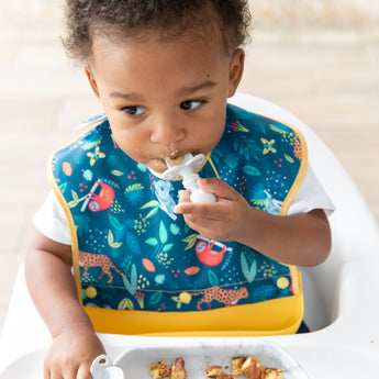 A toddler in a high chair wearing a jungle-themed bib eats from Bumkins Grip Dish Essential Infant 4-Piece Mealtime Set: Marble.