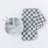 Bumkins Spoon + Fork: Marble sit inside a matching divided plate next to a folded gray & white checkered bib on a white background.