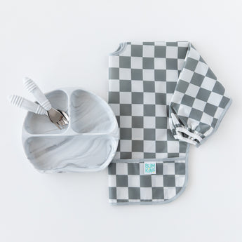 Bumkins Spoon + Fork: Marble sit inside a matching divided plate next to a folded gray & white checkered bib on a white background.