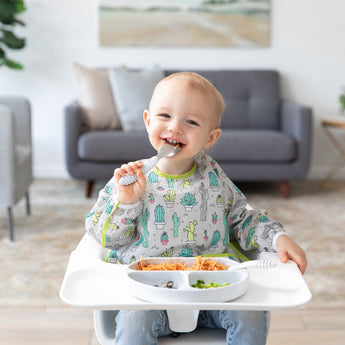 A toddler in a high chair wearing a cactus-print bib enjoys spaghetti with Bumkins Grip Dish Essential Toddler 4-Piece Mealtime Set: Marble.