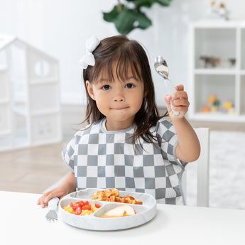 A girl with a bow in a gray & white checkered bib holds Bumkins Spoon + Fork: Marble near a plate with breakfast food on a white table.