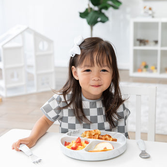 A smiling girl wearing a checkered bib eats breakfast from a Bumkins Silicone Grip Dish: Marble with matching utensils in a bright playroom.