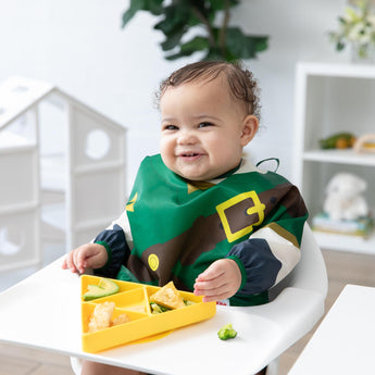 A girl wearing a Bumkins Sleeved Bib: Zelda™ in a high chair smiles in a bright room while eating from a yellow triangle plate.