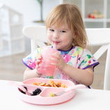 A girl smiles as she drinks from a Bumkins Silicone Straw Cup with Lid: Pink; a pink plate and utensils sit on the table in front of her.