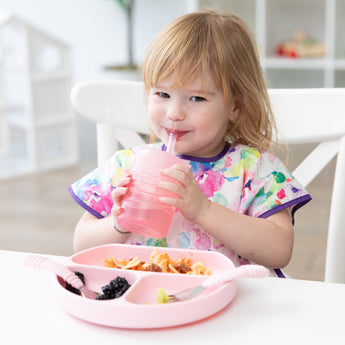 A girl smiles as she drinks from a Bumkins Silicone Straw Cup with Lid: Pink; a pink plate and utensils sit on the table in front of her.