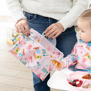 A mom and baby enjoy mealtime with Bumkins On-The-Go Mealtime 6-Piece Set: Boots + Pink in a playroom.