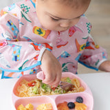 Close-up of a child in a pink patterned bib eating a meal from a Bumkins Silicone Grip Dish: Pink with a pink fork.