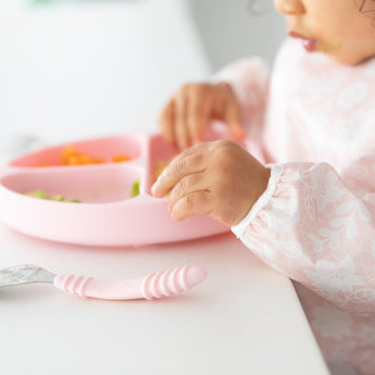Close-up of a child in a pink bib eating from Bumkins Grip Dish Essential Toddler 4-Piece Mealtime Set: Pink at a white table.