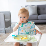 A boy with bright eyes eats breakfast in a high chair from a green plate, wearing a Bumkins SuperBib® 3 Pack: Hangry, Dinosaurs, Blue Tropic.