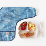 Bumkins Sleeved Bib: Vintage Denim lays flat next to a plate with berries, eggs, & waffles, and toddler utensils on a white background.