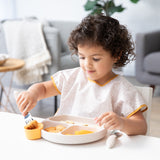 A young child wearing a beige bib eagerly eats from Bumkins Silicone Grip Dish: Sand at a white table in a cozy living room.