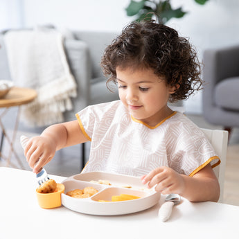 A young child wearing a beige bib eagerly eats from Bumkins Silicone Grip Dish: Sand at a white table in a cozy living room.