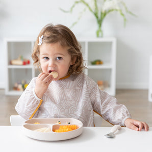 A young girl eats a meal with Bumkins On-The-Go Mealtime 6-Piece Set: Wander + Sand at a white table in a bright playroom.
