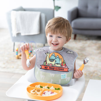 A boy in a high chair eats from a Bumkins Silicone Grip Dish: Tangerine with orange utensils, smiling while wearing a llama themed bib.