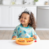 A smiling girl with a bow wearing a blue bib eats from a Bumkins Silicone Grip Dish: Tangerine with matching utensils in a kitchen.