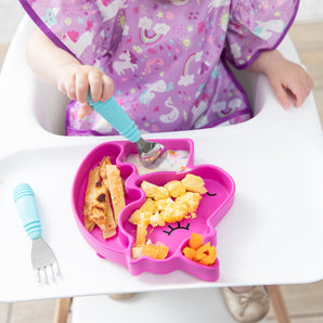 Close-up of a child in a purple patterned bib eating breakfast from a Bumkins Silicone Grip Dish Special Edition: Unicorn with blue utensils.