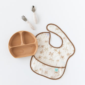 Bumkins Silicone Grip Dish: Wood Grain sits on a white background next to beige utensils and a beige & brown bib on a white background.