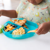 A child in a colorful bib enjoys mac and cheese, fruit, and pizza from a Bumkins Silicone Grip Dish: Blue Jelly at a white table.