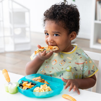 A toddler enjoys pizza at a white table in a bib with fruit, veggies, and pasta in a Bumkins Silicone Grip Dish: Blue Jelly in a bright room.