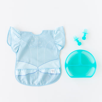 Bumkins Ruffle Apron Bib: Cinderella rests next to a blue divided plate and blue toddler utensils on a white background.