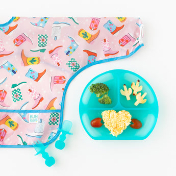 Bumkins Sleeved Bib: Boots lays flat next to a blue divided plate with veggies and rice and matching utensils on a white background.