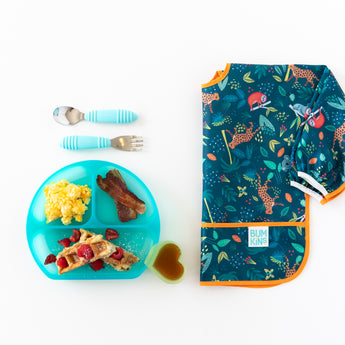 Scrambled eggs, bacon, and waffles in a Bumkins Silicone Grip Dish: Blue Jelly, with cutlery and bright bib nearby on a white background.